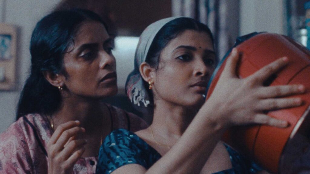 A still from the film "All We Imagine As Light." A closeup of two elegant women, shot in grainy shadow. One holds a modern cooking pot, while the other looks on.