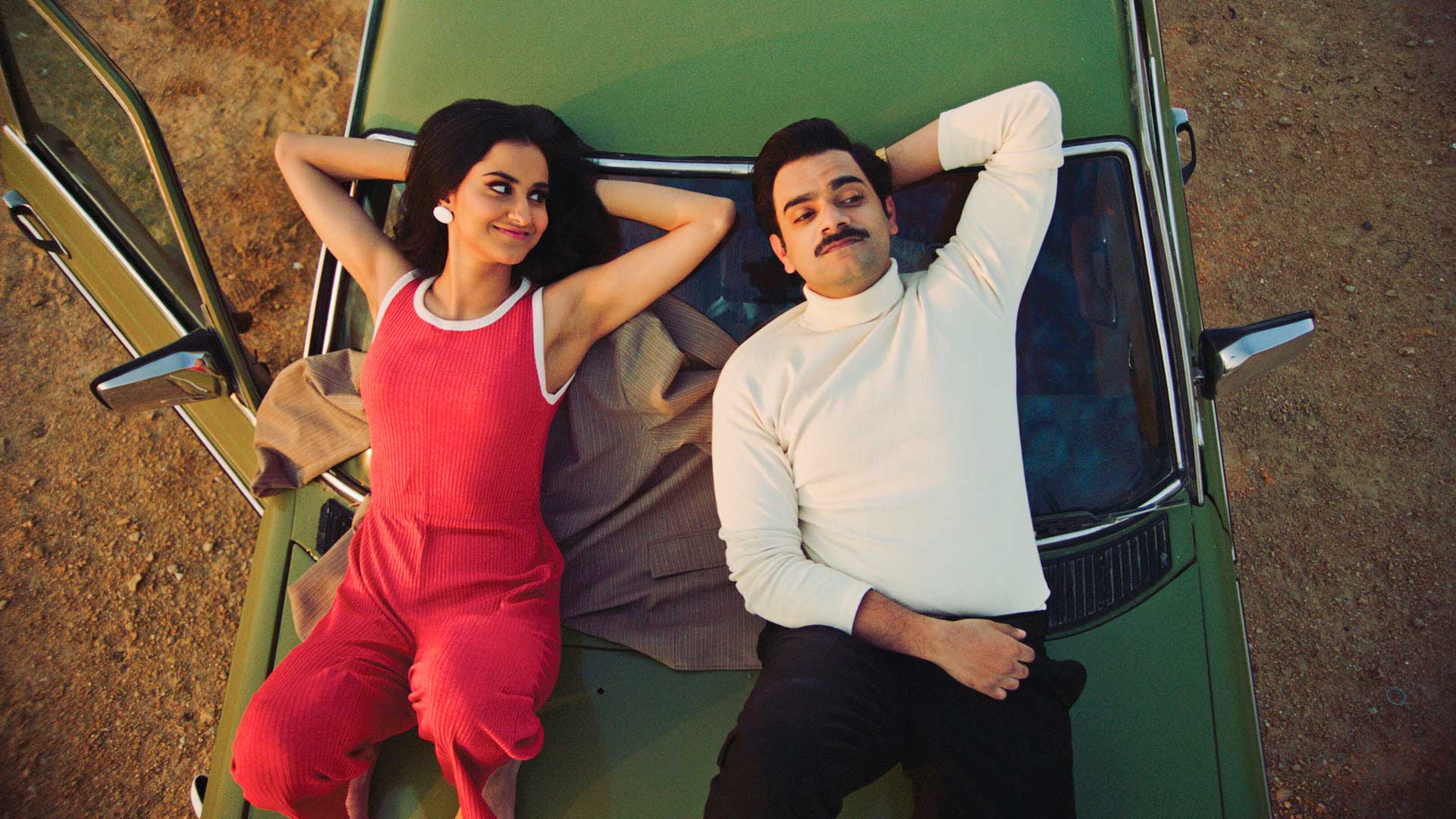 A still from the film, "The Queen of My Dreams." A young South Asian couple leans back on the hood of a car, seen from above. She is wearing a vibrant red jumpsuit and a mischievous expression. He wears a cream-colored turtleneck and a large moustache, and seems pensive but amused.