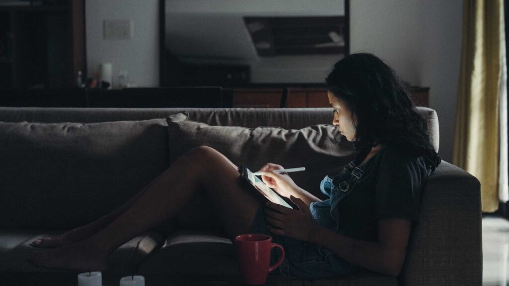 Indian cartoonist Rachita Taneja works on an electronic tablet while seated on a couch. The room is dark, and her face is lit by the tablet.