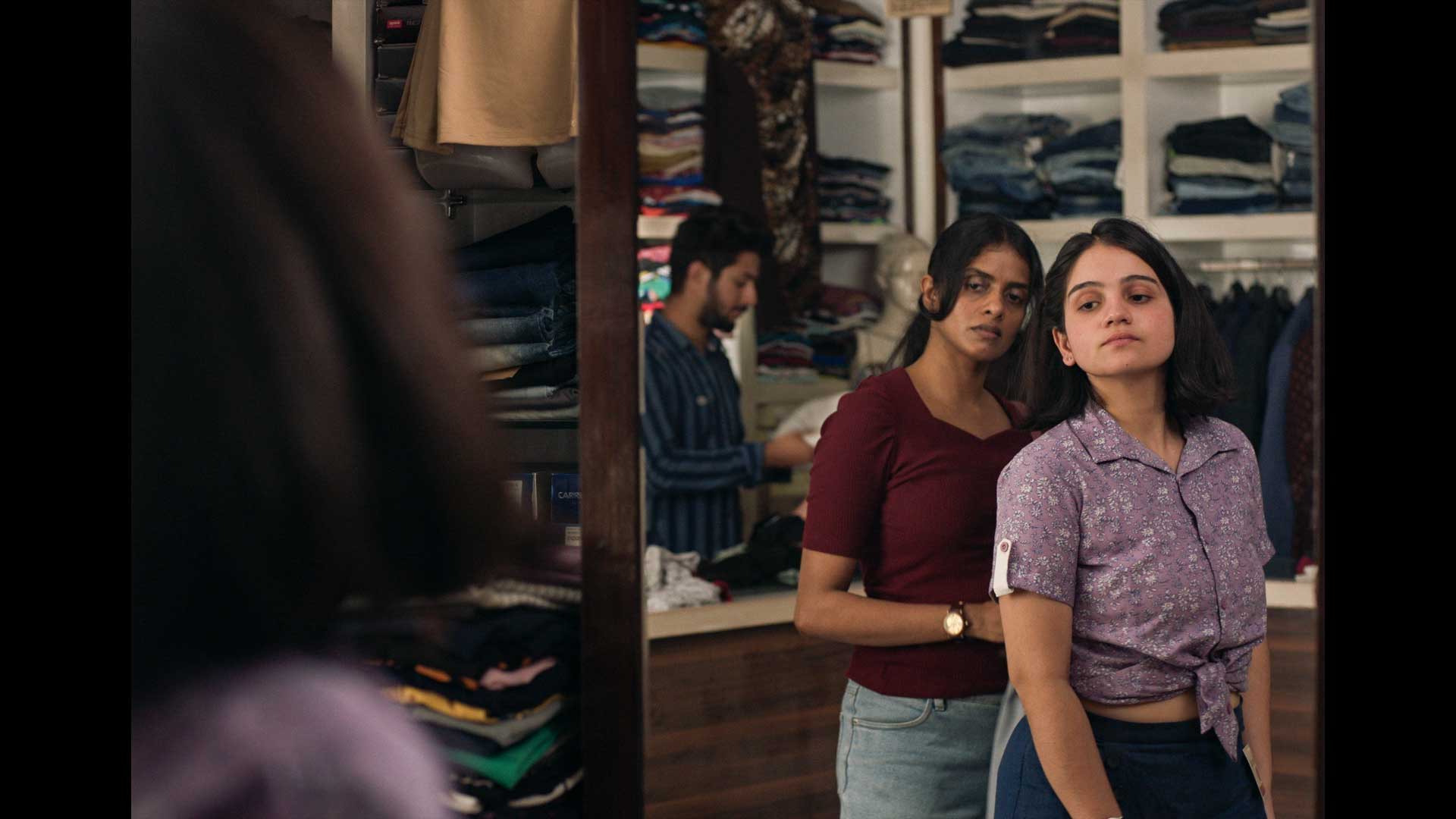 A young woman tries on clothing in a shop, while an older woman looks on behind her. Both are viewing themselves in a mirror, with shelves of clothing behind them.