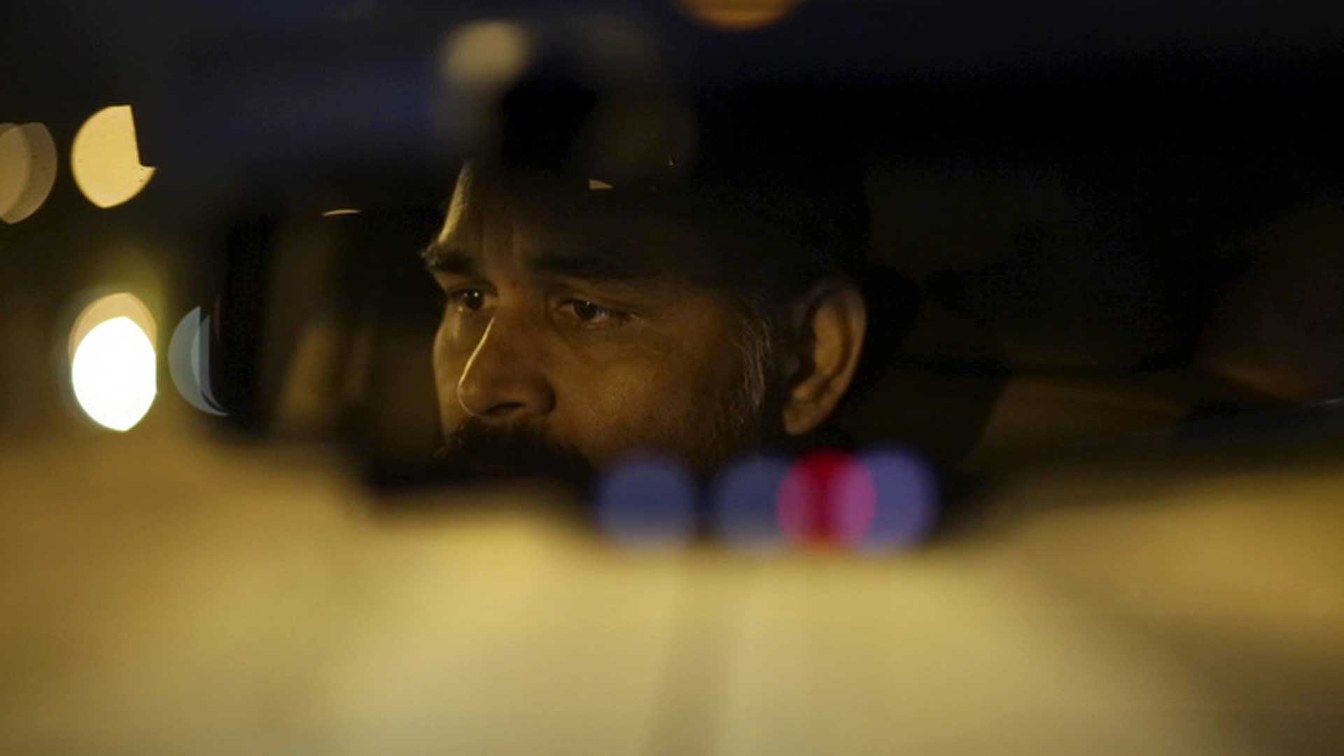 A South Asian man is viewed through the rear-view mirror of a taxi he is driving, at night. It's a moody, semi-abstract shot with reflected lights. Film still from "Land of Dreams," 2024.