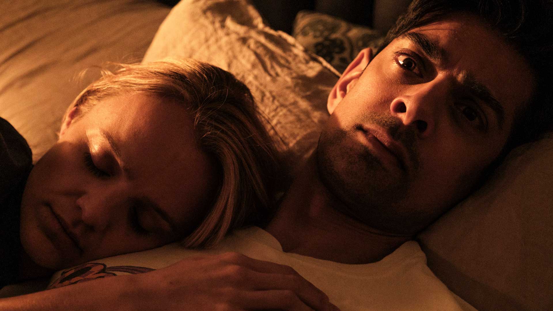 Still from the short film, "Tall Dark and Handsome" (dir. Sam Baron, UK, 2023) An amber-lit closeup of a young blond woman and a young South Asian man's faces, as they lay in a bed. The woman appears to be sleeping peacefully. The man is wide-eyed and appears agitated.