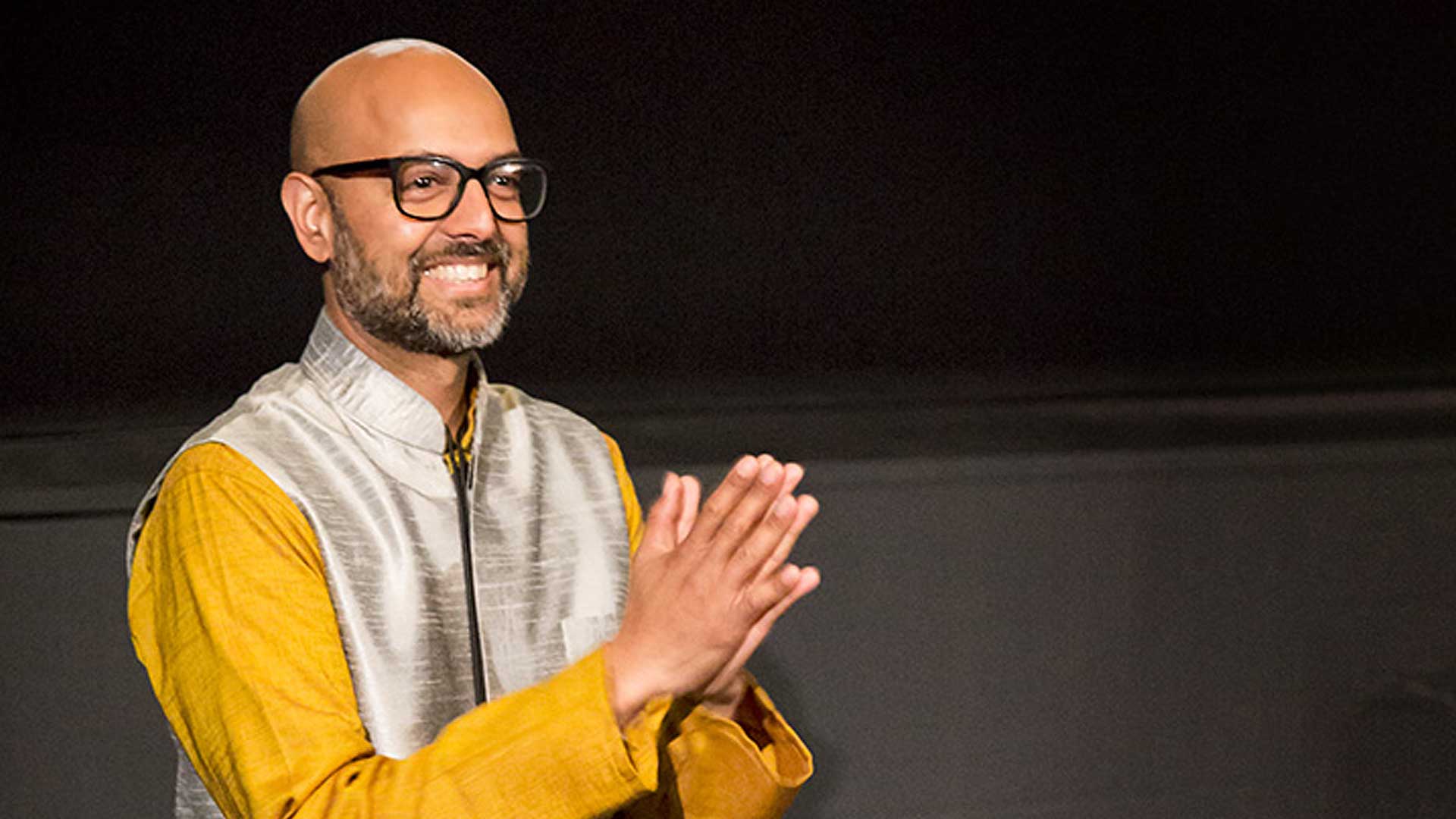 Picture of Robin Sukhadia, a stylish man in big fashionable glasses, wearing a saffron shirt and a silver Nehru-style vest/jacket.